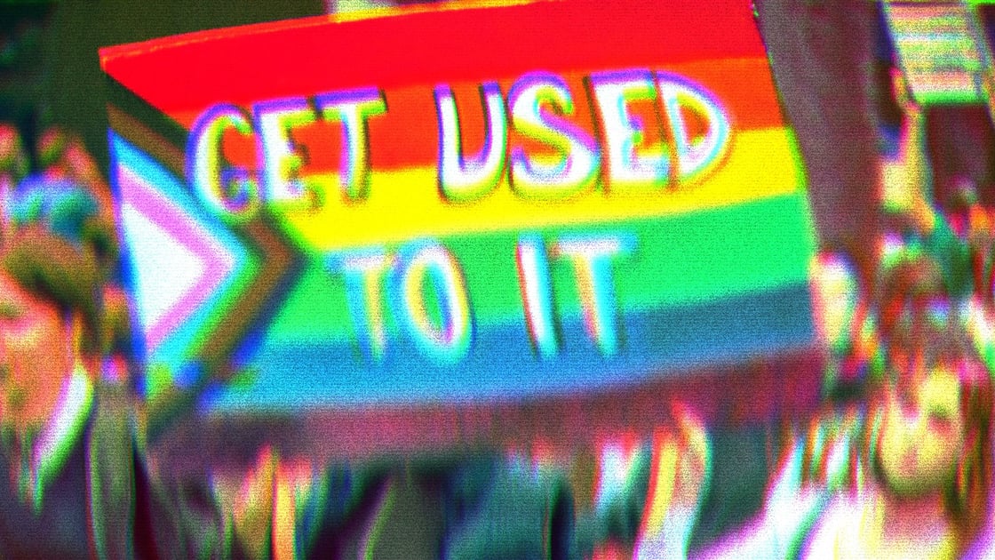 Two people holding a sign that says Get Used To It. The sign background is the modern pride flag